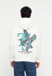 Hvit hettegenser med hette som har trykt duck-motiv, stor "TJ" i teal, og teksten "TOMMY JEANS New York City". Mykt, bomullsmateriale.