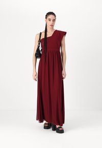Rochie maxi burgundy cu design cu un umăr, material fluid, talie strânsă și bust texturat. Asortată cu sandale cu platformă și o geantă neagră.