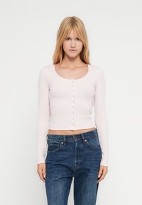 Ξανθιά γυναίκα με ίσια μαλλιά φοράει ένα ανοιχτό ροζ μακρυμάνικο κουμπωτό κοντό τοπ και ψηλόμεσα μπλε τζιν, στέκεται μπροστά σε λευκό φόντο.