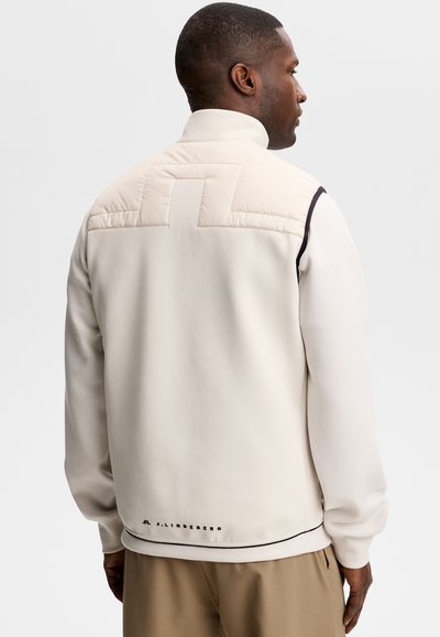 Uomo di spalle che indossa una giacca color crema con dettaglio trapuntato sulla parte superiore della schiena e pantaloni color cuoio, su sfondo semplice grigio chiaro.