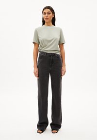 Hellgrünes kurzärmliges T-Shirt, kombiniert mit hochtaillierten schwarzen weiten Jeans. Das Modell trägt schwarze Sandalen mit offenen Zehen. Einfaches, minimalistisches Design.