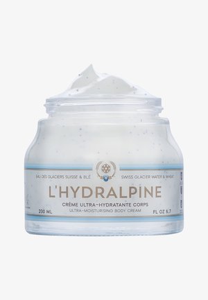 Die ultra-feuchtigkeitsspendende Körpercreme L'Hydralpine in einem klaren Glasbehälter. Cremige weiße Textur mit blauen Sprenkeln. Das Etikett ist mit hellblauen und goldenen Akzenten versehen.