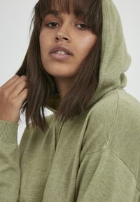 Sudadera color verde oliva hecha de un tejido texturizado, que cuenta con un bolsillo frontal y una capucha grande. Modelo con el cabello largo está parcialmente visible.