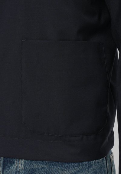 Giacca blu navy con una grande tasca quadrata, texture liscia e cuciture a contrasto lungo l'orlo e il bordo della tasca.