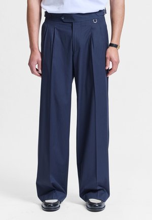 Marineblå stripete vide bukser med frontfolder, knapp og sideregulering, brukt med svart-hvite sko og en hvit skjorte.