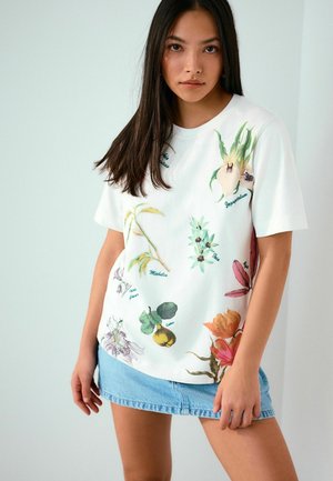 Young woman with long dark hair wearing a white botanical print t-shirt and a light blue denim skirt standing against a plain light background.