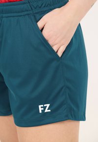 Hand som vilar i sidofickan på turkosa träningstights med "FZ"-logotypen på nedre vänstra benet.