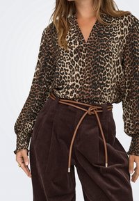 Femme portant un chemisier à imprimé léopard avec des poignets froncés, rentré dans un pantalon en velours côtelé marron foncé taille haute, cintré avec une fine ceinture en cuir nouée.