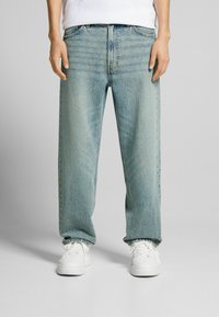 Persona vestida con jeans de pierna ancha color azul claro y zapatillas blancas, de pie contra un fondo gris liso.