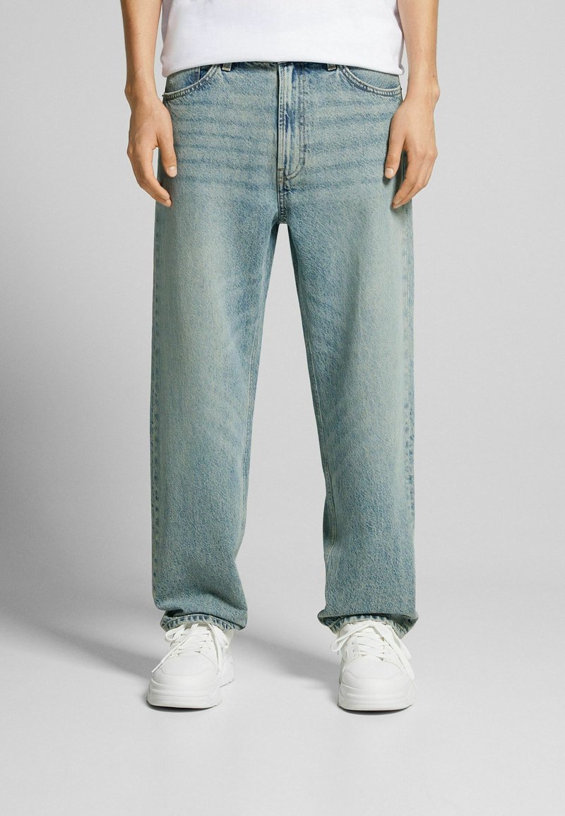 Persona vestida con jeans de pierna ancha color azul claro y zapatillas blancas, de pie contra un fondo gris liso.