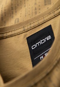 Brun bomulds t-shirt med rund halsudskæring, med et sort mærke med "ombre" logo og størrelsesinformation. Diskret mønstret tekstur i stoffet.