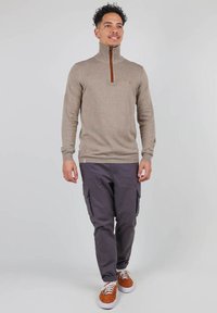 Pull à col zippé beige avec un col côtelé, associé à un pantalon cargo noir. Le mannequin porte des baskets marron avec des semelles blanches.