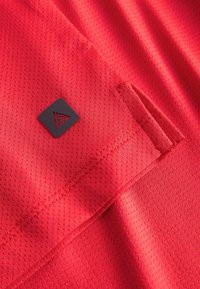 Tissu rouge texturé avec un gros plan sur une couture de poche et un petit logo carré noir fixé près du bord.