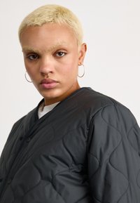 Juoda pasiūta striukė su lygia tekstūra, turinti apvalius mygtukus ir paprastą kaklą. Modelis turi trumpus šviesius plaukus ir žiedines auskarus.