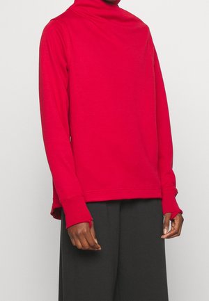Person iført en lys rød langærmet rullekravebluse med tommelfingerhuller på ærmerne og løse sorte bukser mod en ensfarvet baggrund.