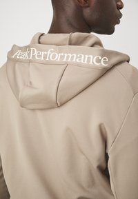 Bežinė gobtuvinė maikutė su dideliu, baltu "Peak Performance" logotipu nugaroje, klasikinio gobtuvo dizainu ir glotnia, lengva medžiagos tekstūra.