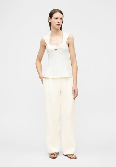 Γυναίκα που φοράει άσπρο αμάνικο τοπ με λεπτομέρεια φιόγκου, κρεμ παντελόνι με φαρδιά πόδια και καφέ σανδάλια, στέκεται με το ένα χέρι στην τσέπη.