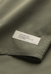 Gros plan sur un tissu vert olive avec une étiquette blanche cousue portant l’inscription OM8RE STREET STYLE since 1985 près de l’ourlet.