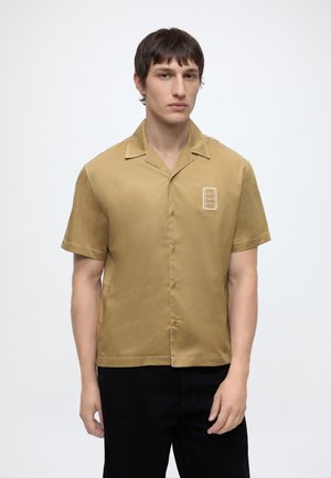 Jeune homme aux cheveux courts et foncés portant une chemise marron clair à manches courtes avec un petit écusson sur la poitrine et un pantalon noir.