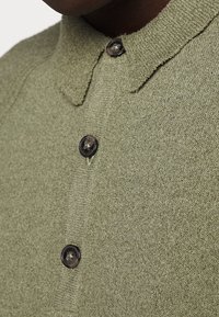 Zelená pletená košile s skládacím límečkem, se černými knoflíky. Materiál má jemnou texturu a ukazuje jemný vzor.