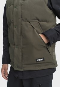 Grüne gepolsterte Weste mit Druckknöpfen, Seitentaschen und einem Markenetikett am Saum. Verfügt über eine glatte Textur und ein warmes Design.