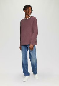 Weinrote Langarmshirt mit einem weißen Rundhalsausschnitt, kombiniert mit hellblauen gerade geschnittenen Jeans und weißen Sneakern. Einfaches Design, lässige Passform.