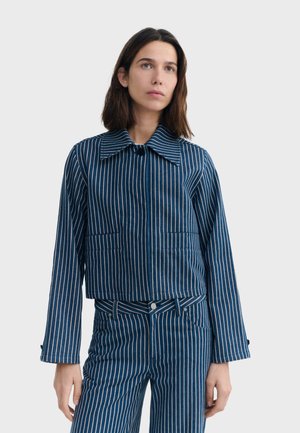 Žena s hnedými vlasmi po plecia, oblečená v zodpovedajúcom modro-bielom pruhovanom saku a nohaviciach, stojaca pred jednoduchým pozadím.