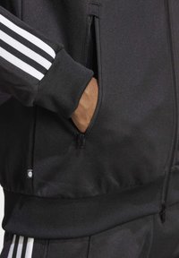 Ruka v kapsičce na zip černé běžecké bundy s bílými pruhy na rukávu, v kombinaci s odpovídajícími černými kalhotami s bílými pruhy.