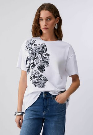 Junge Frau trägt ein weißes T-Shirt mit schwarzem tropischem Blattmuster auf der linken Seite und blaue Jeans, posiert mit der Hand in der Tasche.