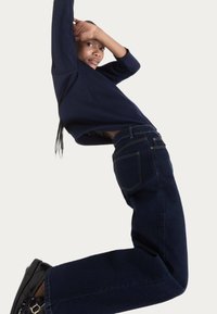 Marine langærmet top parret med højtaljede mørke denimjeans, som har baglommer og synlige sømkanter. Sorte sko fuldender looket.