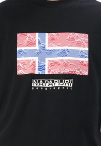 Juodas marškinėlis su dideliu Norvegijos vėliavos grafiku, su banguotomis raudonomis ir mėlynomis linijomis, ir tekstu "NAPAPIJRI geographic" apačioje.