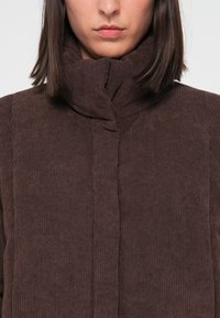 Donna che indossa una giacca in velluto a coste marrone scuro con un colletto alto e imbottito, mostrata dal collo fino a metà torsione su uno sfondo semplice.