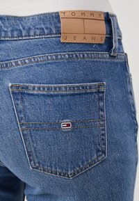 Vaqueros de mezclilla en azul medio con un acabado texturizado. Presenta una etiqueta de cuero marrón con la inscripción "TOMMY JEANS" en la cinturilla y un pequeño logo en el bolsillo trasero.