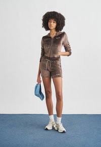 Καφέ βελουτέ μπουφάν με φερμουάρ και κοντό παντελόνι με λευκές πλευρικές ρίγες και κορδόνι, σε συνδυασμό με μπλε καπέλο και ουδέτερα αθλητικά παπούτσια.