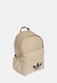 Beżowy plecak z tkaniny z zaokrąglonym górnym zakończeniem, wyposażony w przednią kieszeń i czarne logo. Posiada pionowe paski na ramiączkach.