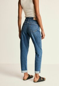 Jean bleu taille haute avec une coupe fuselée, ourlets retroussés et deux poches arrière. Porté avec des sandales noires.
