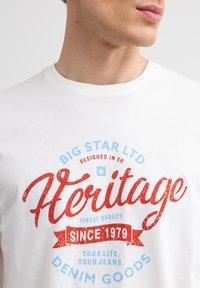 Biała bawełniana koszulka z grafiką w kolorze czerwonym i niebieskim. Tekst zawiera "Big Star LTD", "Heritage" oraz "Since 1979". Klasyczny krój z okrągłym dekoltem.