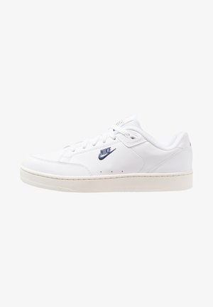 Zapatillas de cuero blancas con una textura suave, que presentan un logo de Nike azul, cordones planos y una suela de goma beige. Diseño minimalista.