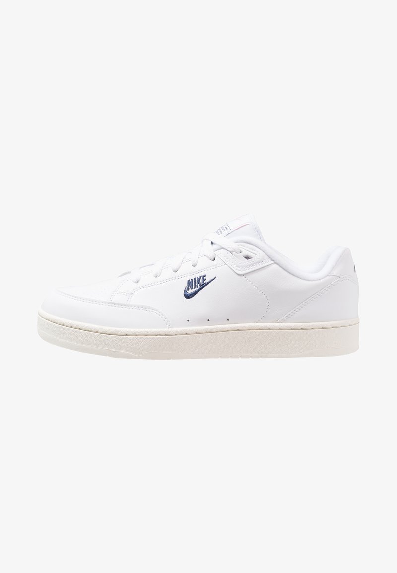 Zapatillas de cuero blancas con una textura suave, que presentan un logo de Nike azul, cordones planos y una suela de goma beige. Diseño minimalista.