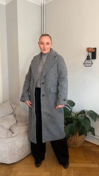Grå ullrock med texturerat tyg, stora slag och knappar, kombinerad med en grå tröja och svarta byxor. Inomhusmiljö med växter.