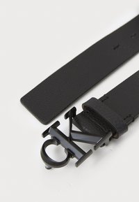 Schwarzer Ledergürtel mit strukturiertem Finish und einer schwarzen Metallschnalle, die die Initialen "CK" in einem geometrischen Design zeigt.