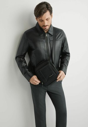 Mężczyzna w czarnej skórzanej kurtce i ciemnych spodniach, dostosowujący czarną torbę na ramię, stojąc na tle białej ściany.