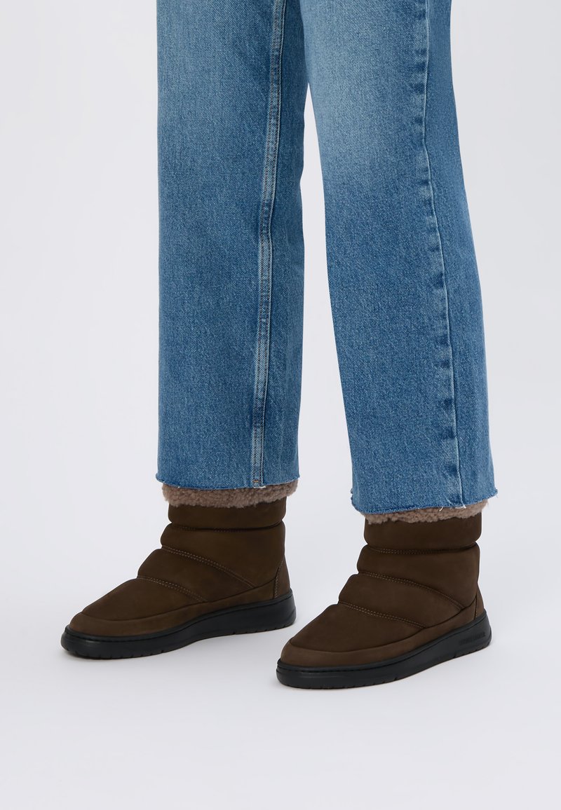 Stivaletti in suede marrone con profilo basso, presentano una texture trapuntata, una fodera morbida e una suola in gomma nera, abbinati a jeans di denim blu con risvolto.