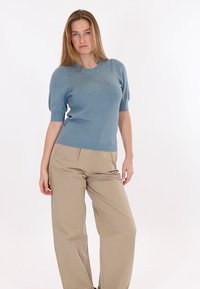 Moteris stovinti viena ranka ant klubo, apsirengusi tekstūruotu mėlynu trumpomis rankovėmis megztiniu ir smėlio spalvos platėjančiomis kelnėmis, stovinti prieš baltą foną.