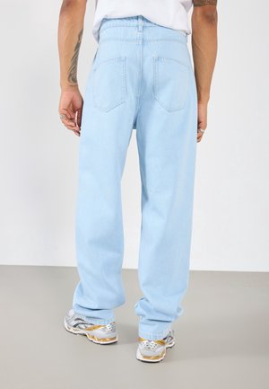 Person wearing light blue loose-fit jeans and silver sneakers, shown from the back against a plain gray and white background.