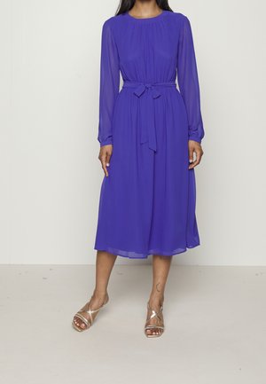 Femme portant une robe violette à manches longues, longueur genoux, avec une ceinture nouée à la taille et des sandales argentées à brides, debout devant un fond uni.