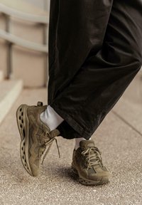 Persona con zapatillas deportivas marrones y calcetines blancos, vestida con pantalones anchos negros, de pie sobre un pavimento texturizado cerca de unas escaleras.