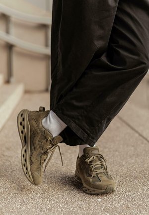 Une personne portant des chaussures de sport marron et des chaussettes blanches, en pantalon large noir, se tient sur un pavé texturé près d'un escalier.