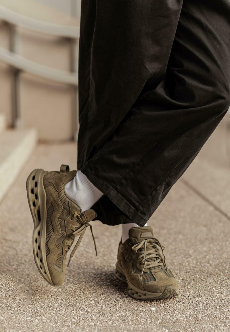 Persona con zapatillas deportivas marrones y calcetines blancos, vestida con pantalones anchos negros, de pie sobre un pavimento texturizado cerca de unas escaleras.