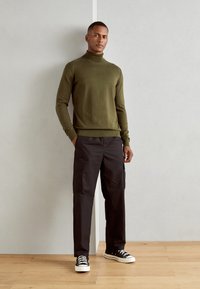 Maglione a collo alto verde oliva, pantaloni cargo neri con tasche laterali e sneaker nere. Texture liscia, design aderente. Pavimento in legno, sfondo neutro.